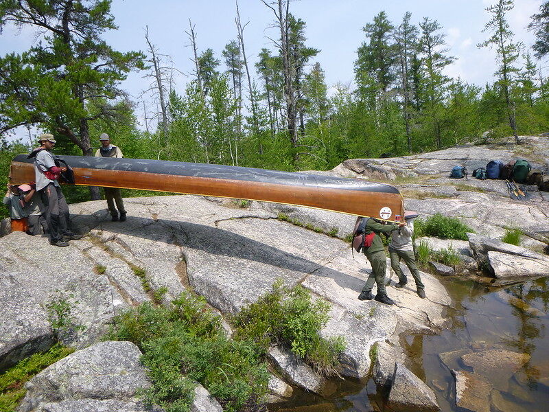 A group of people are carrying a canoe over a rocky terrain near a body of water. The canoe is long and appears heavy, requiring multiple people to lift and transport it. The surrounding environment includes trees and rocks, suggesting a wilderness or outdoor setting. Backpacks are scattered nearby, indicating that the group may be on a camping or hiking trip.